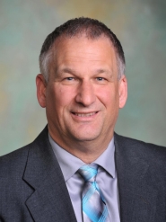 Official photo of Jeff Larshus, Interim Deputy Director. Jeff is a white male with salt and pepper grey hair. He is wearing a grey business suit, dress shirt and tie.