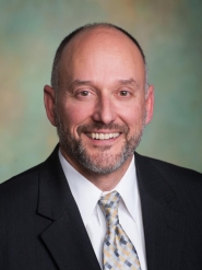 Official photo of Joe Morrissette who is a white male with a beard and mustache wearing a dress shirt, tie, and suit coat.